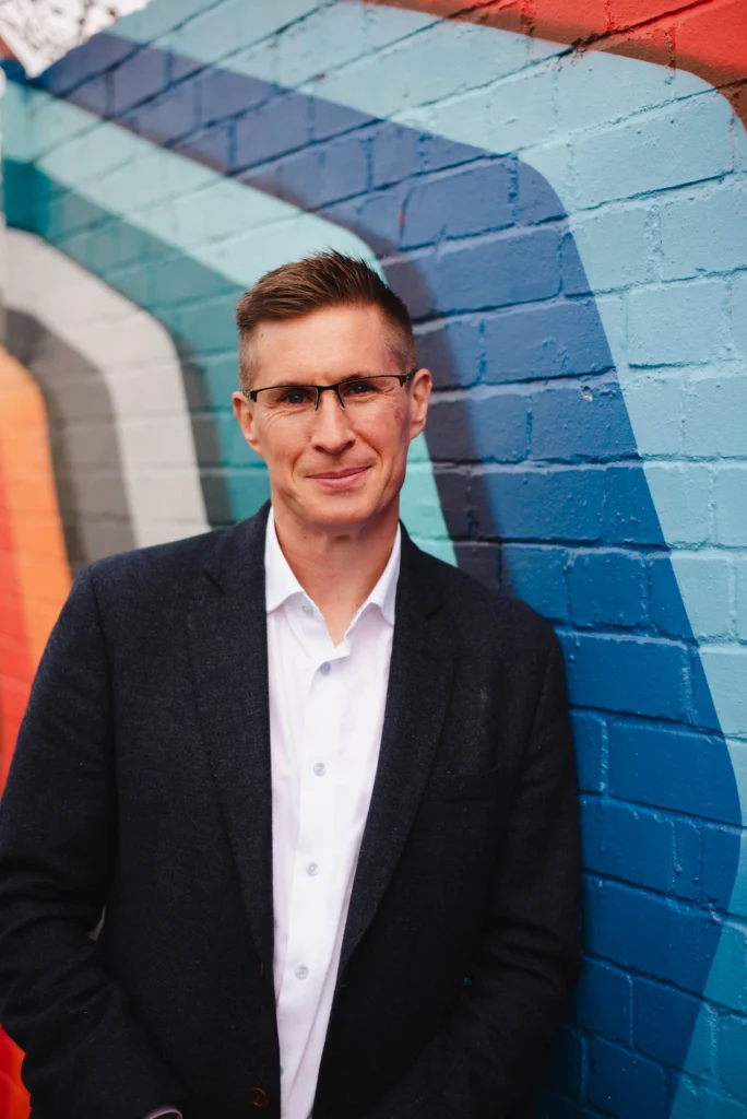 Man in a dark blazer and white shirt stands smiling against a colorful, geometric mural on a brick wall. The mood is casual and approachable.