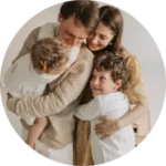 A family of four embraces warmly, each wearing light-colored clothes. The parents hold their children close, smiling, in a tender, loving moment.