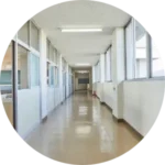 Wide hallway with windows on the right, white walls, and fluorescent lights. Doors line the left side, creating a calm, orderly, institutional feel.