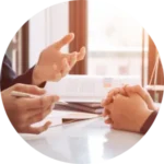 Two people engage in conversation at a desk. One gestures with an open hand while the other folds hands, with documents and sunlight in the background.